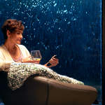 A woman relaxing in a chair with a glass of wine while using a smartphone to control smart home features, with illuminated water cascading down a glass wall behind her.